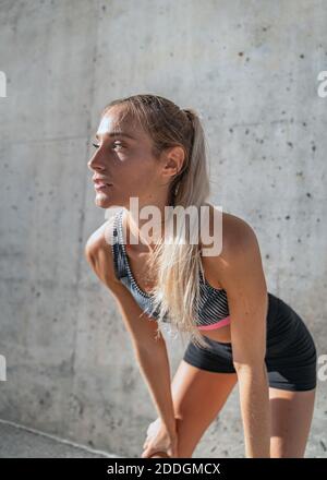 Athlète féminine fatiguée en vêtements d'activité se pliant vers l'avant, debout près du béton repos mural pendant l'entraînement en ville Banque D'Images