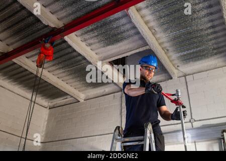 Faible angle d'homme joyeux ouvrier en casque de sécurité et de protection lunettes se tenant sur l'échelle et en installant un tuyau métallique avec du plombier clé lors de l'intervention Banque D'Images