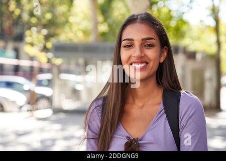 Agréable jeune ethnique long cheveux femme en tenue décontractée avec lunettes de soleil souriant et regardant l'appareil photo tout en se tenant contre flou environnement urbain Banque D'Images