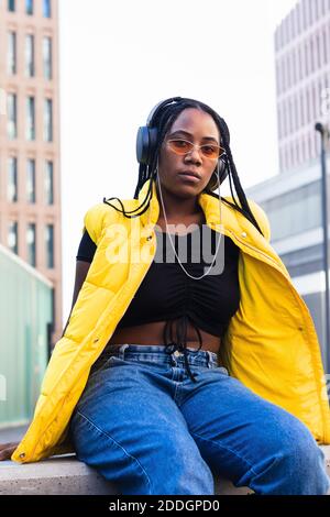 Jeune femme noire en gilet tendance et lunettes de soleil avec casque et profiter de votre musique préférée en regardant votre appareil photo dans le centre-ville Banque D'Images