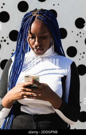 Déterminée afro-américaine avec des tresses bleues debout en milieu urbain environnement utilisant un smartphone Banque D'Images