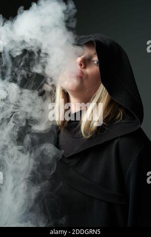 Rebelle jeune femme dans le noir à capuche fume e cigarette et vapeur expiratoire en regardant l'appareil photo sur fond gris foncé Banque D'Images