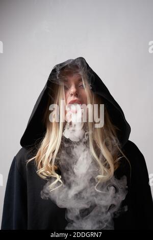 Rebelle jeune femme dans le noir à capuche fume e cigarette et vapeur expiratoire en regardant l'appareil photo sur fond gris foncé Banque D'Images