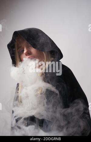 Rebelle jeune femme dans le noir à capuche fume e cigarette et vapeur expiratoire en regardant l'appareil photo sur fond gris foncé Banque D'Images