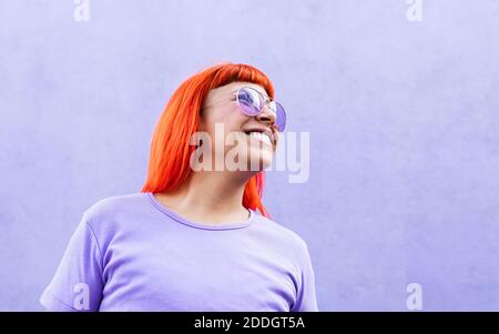 Femme adulte aux cheveux teints rouge vif et élégante lunettes de soleil qui se trouvent à l'écart du mur violet dans la rue Banque D'Images