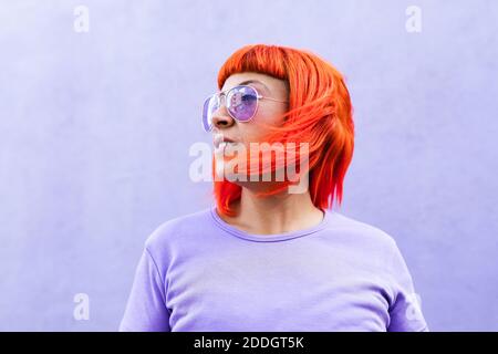 Femme adulte aux cheveux teints rouge vif et élégante lunettes de soleil qui se trouvent à l'écart du mur violet dans la rue Banque D'Images