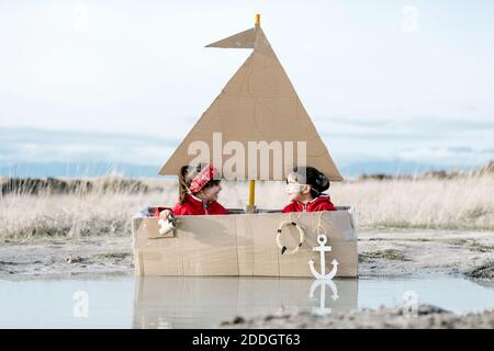 Enfants joueurs assis dans un bateau en carton fait main dans une flaque et regarder à travers le spyglass tout en s'amusant le week-end ensemble Banque D'Images