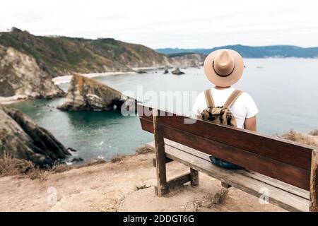 Vue arrière angle élevé de l'explorateur masculin dans le chapeau assis sur un banc en bois et en admirant le paysage spectaculaire de la mer et rochers Banque D'Images