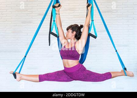 Femme mince faisant des fentes avec les bras relevés sur les silks aériens tout en pratiquant Pilates contre le mur de brique dans la salle de gym Banque D'Images