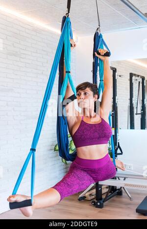 Femme mince faisant des fentes avec les bras relevés sur les silks aériens tout en pratiquant Pilates contre le mur de brique dans la salle de gym Banque D'Images