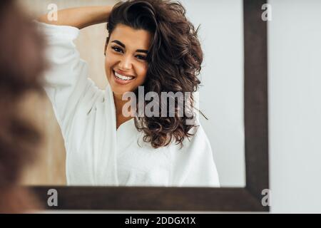 Belle femme souriante portant un peignoir et tenant ses cheveux en vous tenant devant un miroir Banque D'Images
