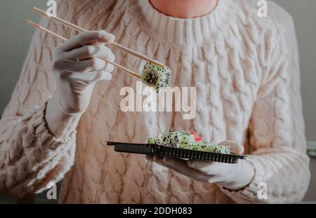 Main dans un gant en caoutchouc tenir les sushis avec un bâton au-dessus d'un contenant jetable de livraison d'aliments. Menu sushi. Cuisine japonaise. Banque D'Images