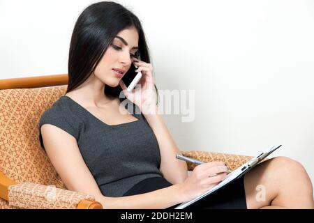 Femme caucasienne assise sur un fauteuil. Elle parle sur le téléphone portable et prend des notes. Banque D'Images