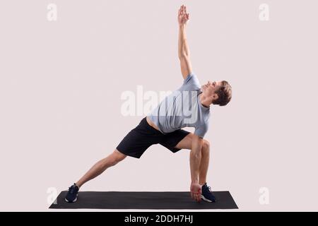 Un jeune homme fort debout dans le yoga pose sur un tapis de sport sur fond léger de studio, portrait en longueur Banque D'Images