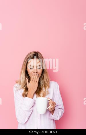 Jeune femme en pyjama bâillant et tenant la tasse sur rose arrière-plan Banque D'Images