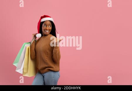 Énorme solde de Noël. Belle femme noire dans le chapeau de Santa tenant des sacs-cadeaux et pointant à l'espace vide, fond rose Banque D'Images