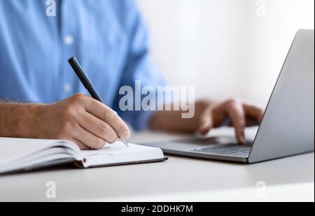 Homme méconnaissable écrivant dans le Bloc-notes et utilisant l'ordinateur portable dans le bureau, gros plan Banque D'Images