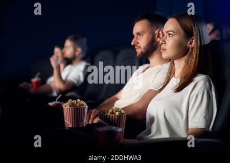Objectif sélectif de jeunes couples regardant un film au cinéma, assis dans des sièges noirs confortables, portant des t-shirts blancs. Vue latérale de l'homme et de la femme caucasiens concentrés avec des visages forts appréciant le film. Banque D'Images