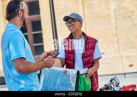 Déjeuner frais. Jeune coursier asiatique joyeux avec sac thermo livrant de la nourriture au client sur le scooter. Concept des services de livraison Banque D'Images