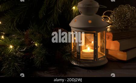 Lanterne blanche avec une bougie allumée. Livres, branche d'arbre de Noël et lumières sur fond. Image sombre de nuit. Humeur mystère de Noël. Banque D'Images