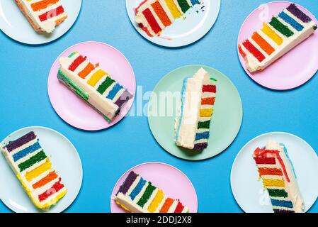 Tranches de gâteau arc-en-ciel sur des assiettes colorées, isolées sur fond bleu. Étaler à plat avec un gâteau à la crème au beurre. Table de fête avec dessert festif. Banque D'Images