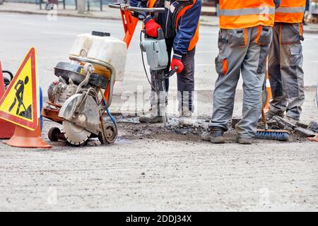 Trois employés d'entretien routier en vêtements réfléchissants sur une section clôturée de la route réparent la route à l'aide d'un outil de route portatif et d'un marteau-piqueur. Banque D'Images