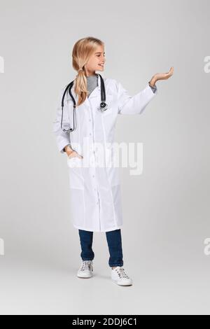 Mignon petite fille d'enfant dans le manteau de docteur avec stéthoscope pointe sur le côté sur fond de studio. Enfant jouant docteur Banque D'Images