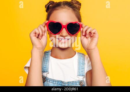 Adolescent élégant avec des verres en forme de coeur, fille heureuse posant sur fond jaune Banque D'Images