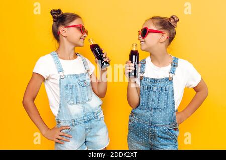 Deux adolescentes, amies élégantes avec lunettes de soleil et boissons non alcoolisées dans les mains, se reposent sur un fond jaune Banque D'Images