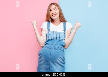 une jeune femme enceinte pensive et heureuse, en combinaison denim, regarde soigneusement et montre un geste de victoire et de succès, debout sur un rose et Banque D'Images