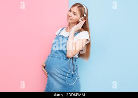 Bonne mignonne belle jeune gaie femme enceinte aime écouter musique avec casque tout en restant sur un rose isolé et arrière-plan bleu Banque D'Images