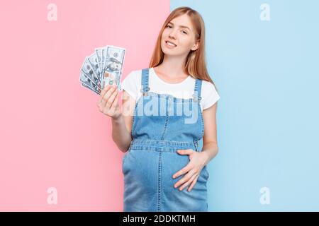 Bonne jeune femme enceinte tenant des billets de dollars, fille, debout sur un fond rose et bleu, le concept de la maternité de substitution Banque D'Images