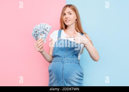 Bonne jeune femme enceinte tenant des billets de dollars, fille, debout sur un fond rose et bleu, le concept de la maternité de substitution Banque D'Images