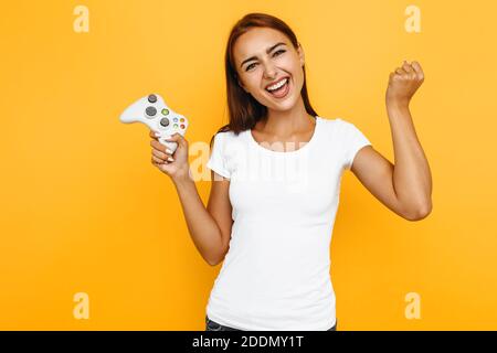 Jeune fille excitée, dans un t-shirt blanc, jouant un jeu vidéo avec un joystick, se réjouit de la victoire, sur un fond jaune Banque D'Images