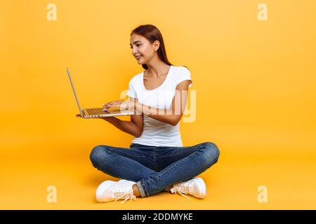 Portrait d'une jeune femme souriante assise à pattes croisées, à l'aide d'un ordinateur portable isolé sur fond jaune Banque D'Images