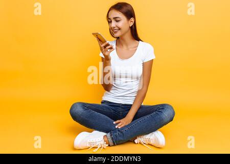 Jeune belle fille, à pattes croisées, sur le sol, envoie un message vocal à un smartphone, sur fond jaune Banque D'Images