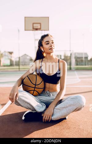 Jeune fille sportive attrayante tenant un basket-ball tout en s'asseyant dans un stade de sport, à l'extérieur. Repos après l'entraînement Banque D'Images