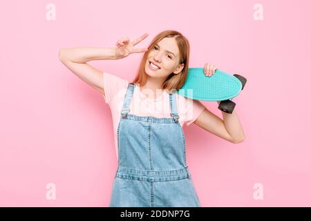 Portrait d'une fille de patineuse attrayante tenant un skateboard et montrant un signe de paix isolé sur un fond rose dans Un studio Banque D'Images