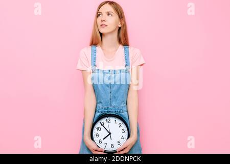Une jeune fille pensive avec une horloge murale dans ses mains, regardant attentivement dans la distance sur un fond rose isolé Banque D'Images