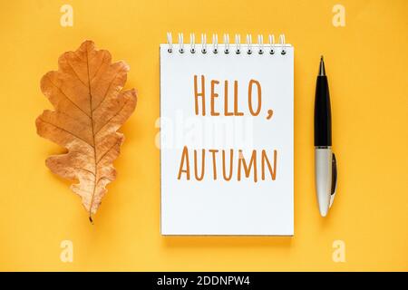 Bonjour automne texte dans bloc-notes, feuille de chêne orange séché et stylo sur fond jaune. Vue du dessus carte de vœux à plat. Banque D'Images