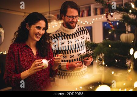 Femme nouant une boule décorative brillante sur l'arbre de Noël avec un homme debout près. Couple décorant arbre de Noël à la maison de famille. Banque D'Images