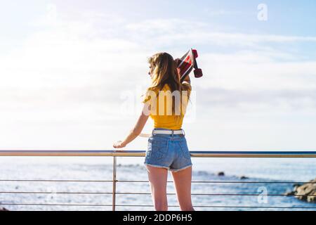 Une jeune femme caucasienne aux cheveux blonds tenant un skateboard et profiter de la vue sur la mer Banque D'Images