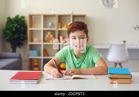 École en ligne. Garçon souriant assis à un bureau avec des carnets et des manuels, faisant des devoirs et regardant la caméra sur l'arrière-plan intérieur de la pièce. Banque D'Images