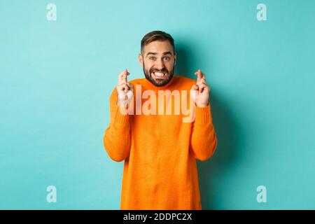 Inquiet homme faisant un souhait, en espérant quelque chose, croiser les doigts bonne chance, se tenant nerveux sur fond turquoise Banque D'Images