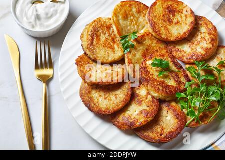 Crêpes de pommes de terre au fromage cottage à l'ail, persil, servies avec une sauce à la crème sure sur une assiette sur fond de pierre en marbre clair avec couverts dorés Banque D'Images