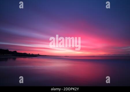 Une longue exposition capture un ciel rempli de nuages roses et violets striés alors que le soleil commence à s'élever au-dessus de la mer. Le sable humide reflète les couleurs vives. Banque D'Images