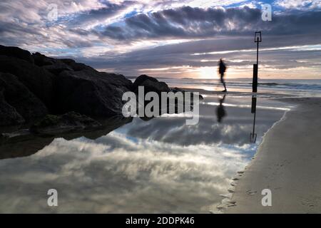 La figure éclipsable et silhouettée d'un homme qui se promène le long de la plage se reflète dans un bassin d'eau de mer calme tandis que le soleil se lève dans un ciel nuageux. Banque D'Images