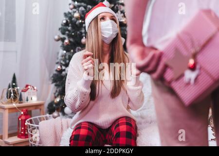 Couple romantique échangeant des cadeaux. Femme dans un masque médical de protection à Noël. Vue arrière rapprochée de l'homme tenant la boîte présente derrière son dos pendant que s Banque D'Images