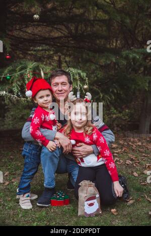 Père en chapeau de Noël rouge et deux filles en chandails rouges heureux près de l'arbre de Noël en plein air dans la cour de la maison avant les vacances, europe Banque D'Images