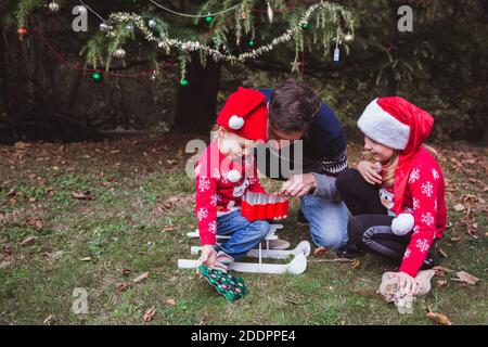 Père et deux filles ouvertes présente des boxes près de l'arbre de Noël en plein air dans la cour de la maison. Joyeux Noël et joyeuses fêtes Banque D'Images